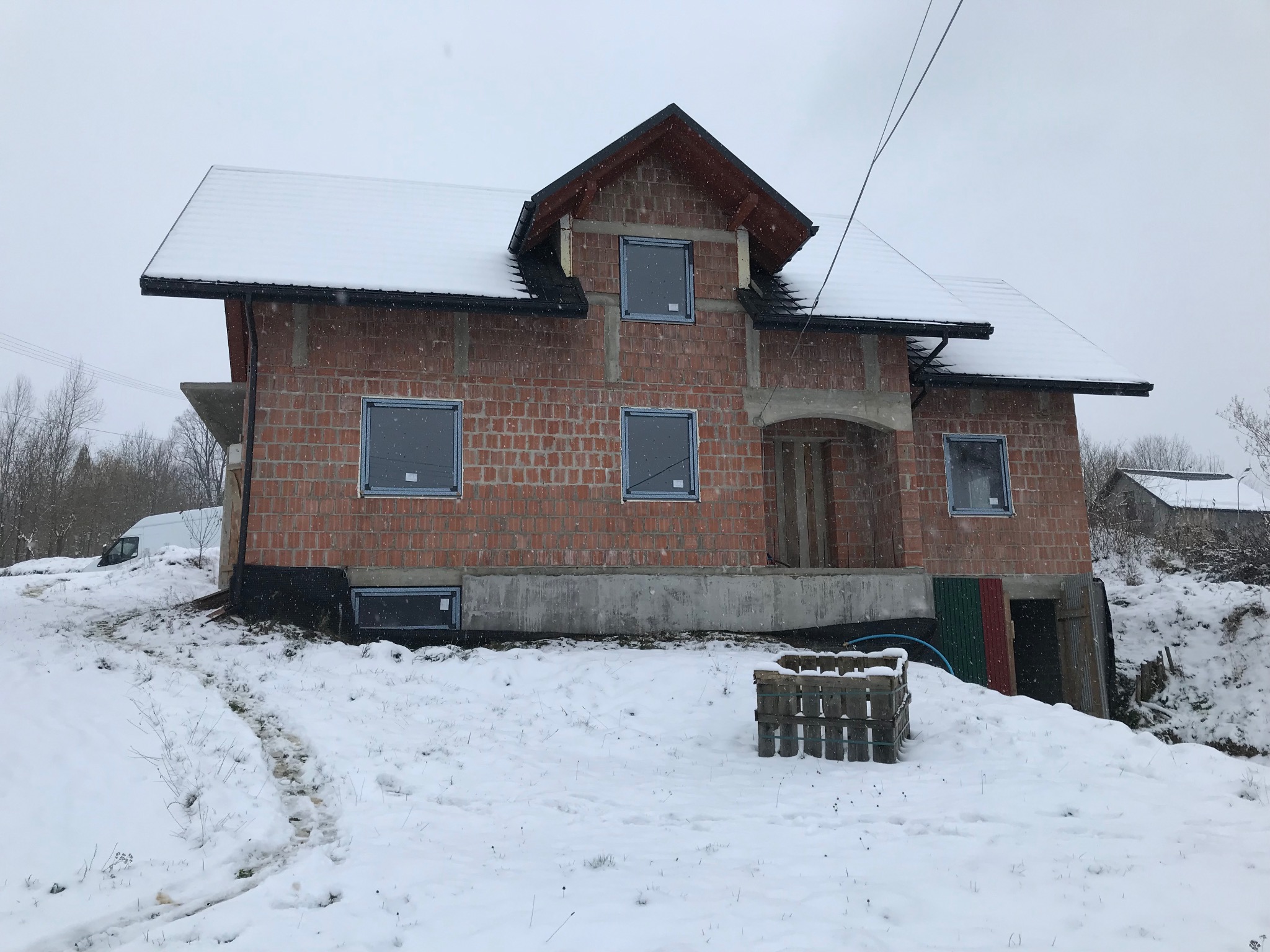 Surowy budynek w trakcie budowy, z zamontowanymi oknami w szarych ramach, pokryty śniegiem, widoczne ślady w śniegu i paleta z pustaków.