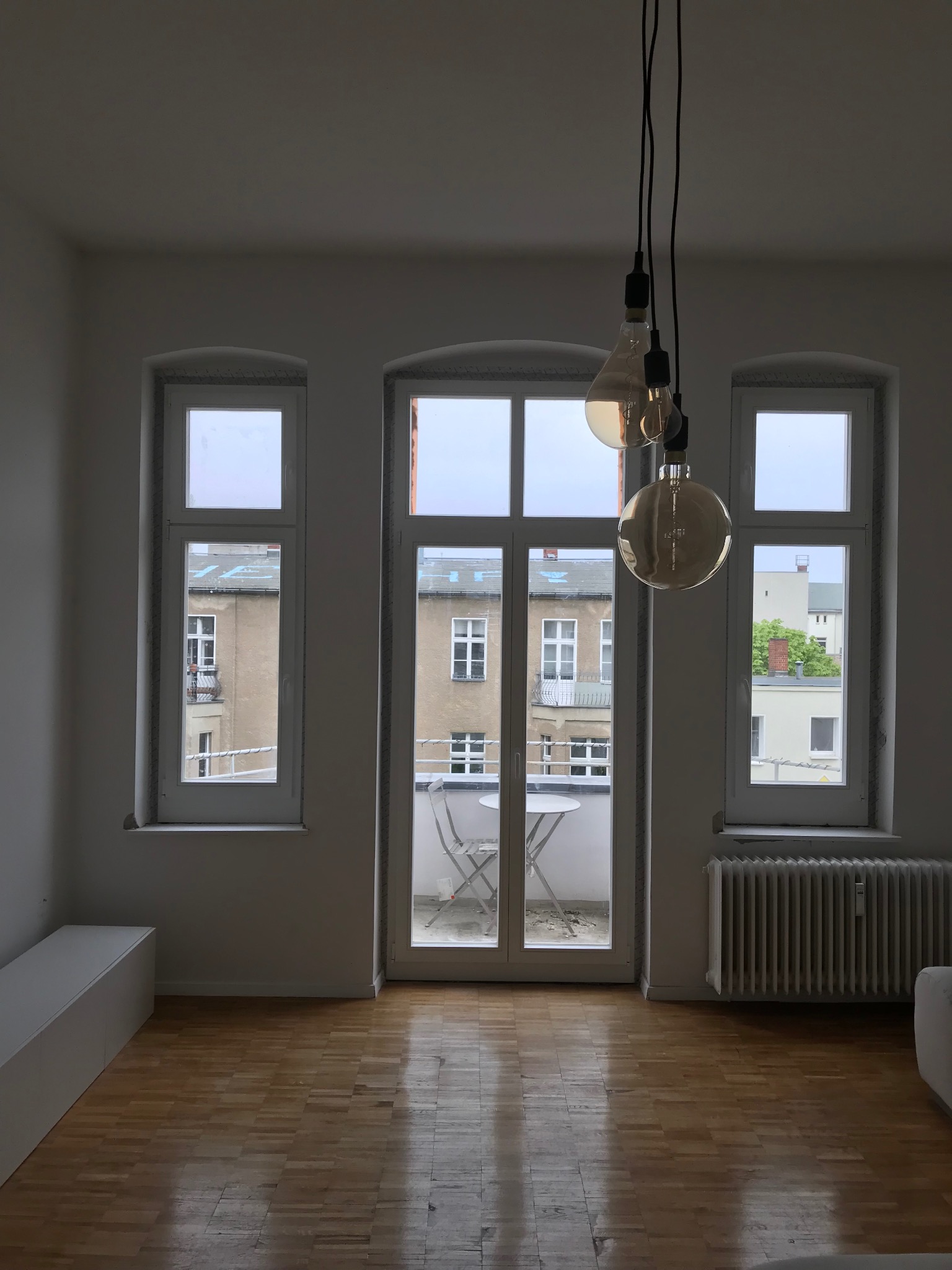 Wnętrze pokoju z trzema oknami z białymi ramami, parkietem i wiszącymi lampami dekoracyjnymi.