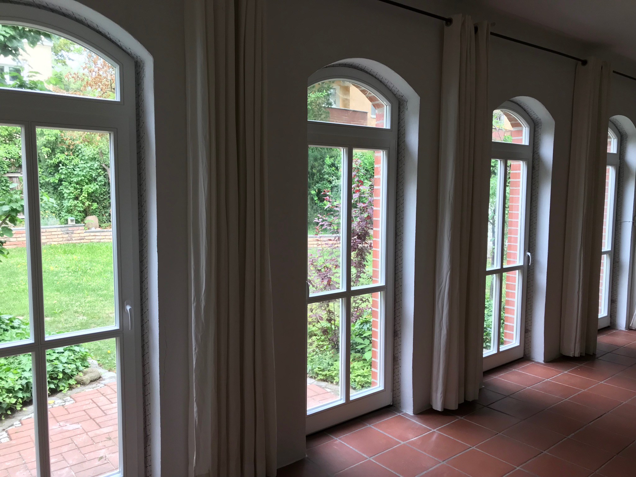 Rząd białych okien z łukowym zwieńczeniem, zasłoniętych kremowymi zasłonami, widok na zielony ogród z ceglanym chodnikiem, czerwona terakota na podłodze.