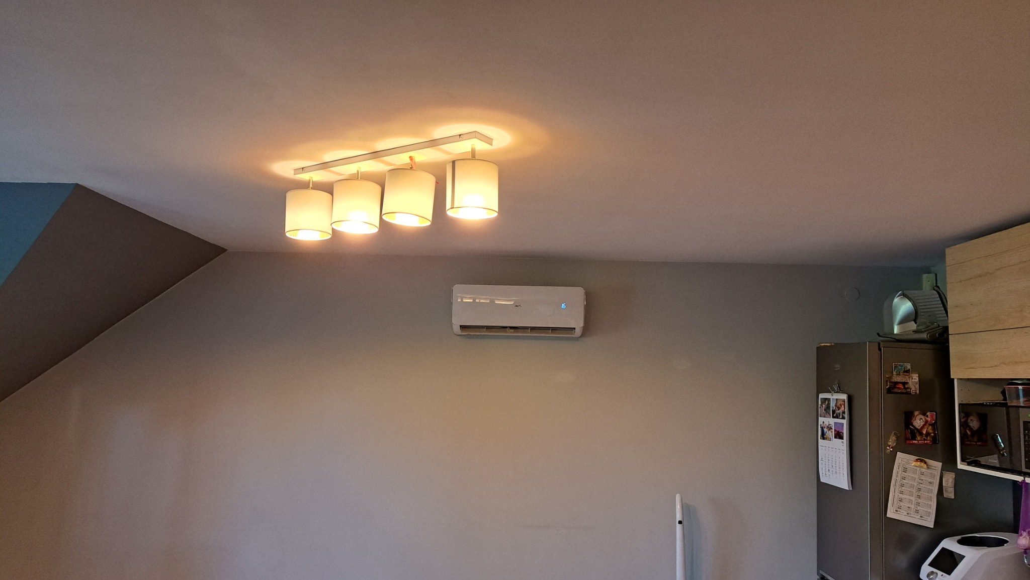 Biały klimatyzator ścienny zamontowany w szarym pokoju pod lampą sufitową z czterema abażurami, widoczny fragment lodówki i zabudowy kuchennej.