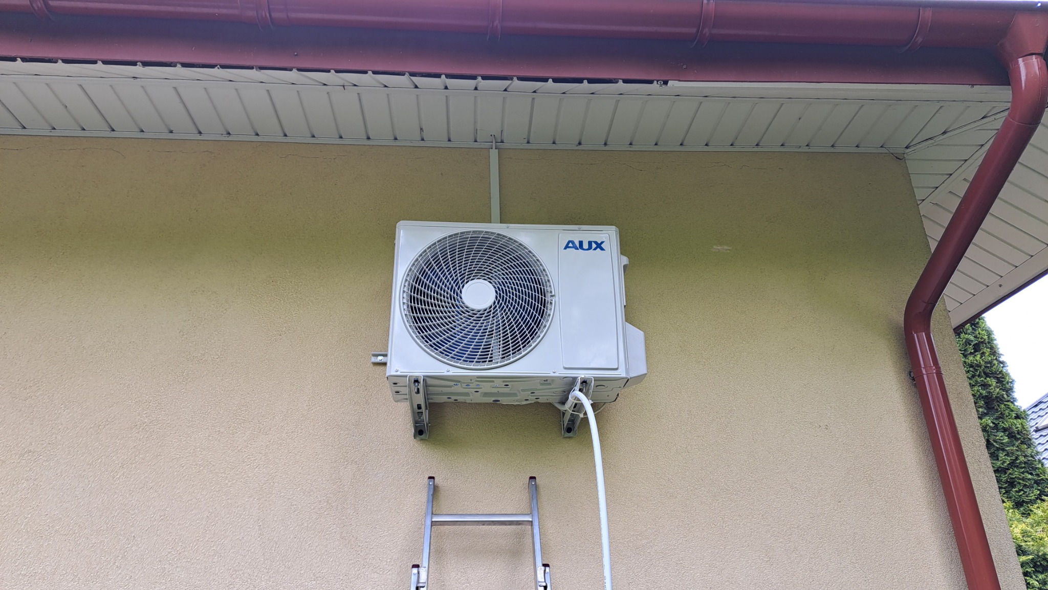 Zewnętrzna jednostka klimatyzacji marki AUX zamontowana na elewacji budynku z widoczną drabinką i rurą odprowadzającą skropliny.