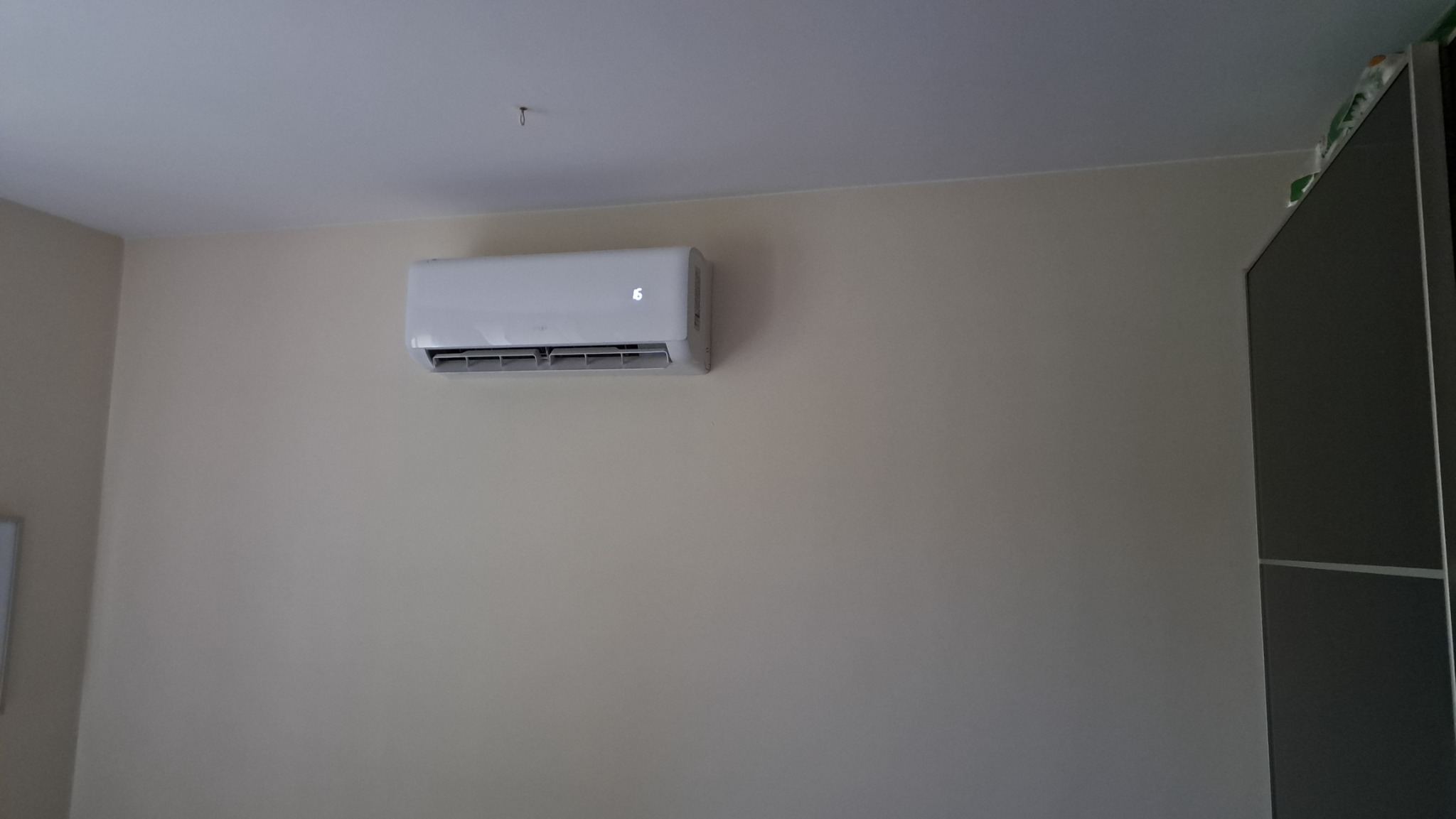 Biały klimatyzator ścienny z wyświetlaczem temperatury '16' zamontowany na beżowej ścianie w pokoju.