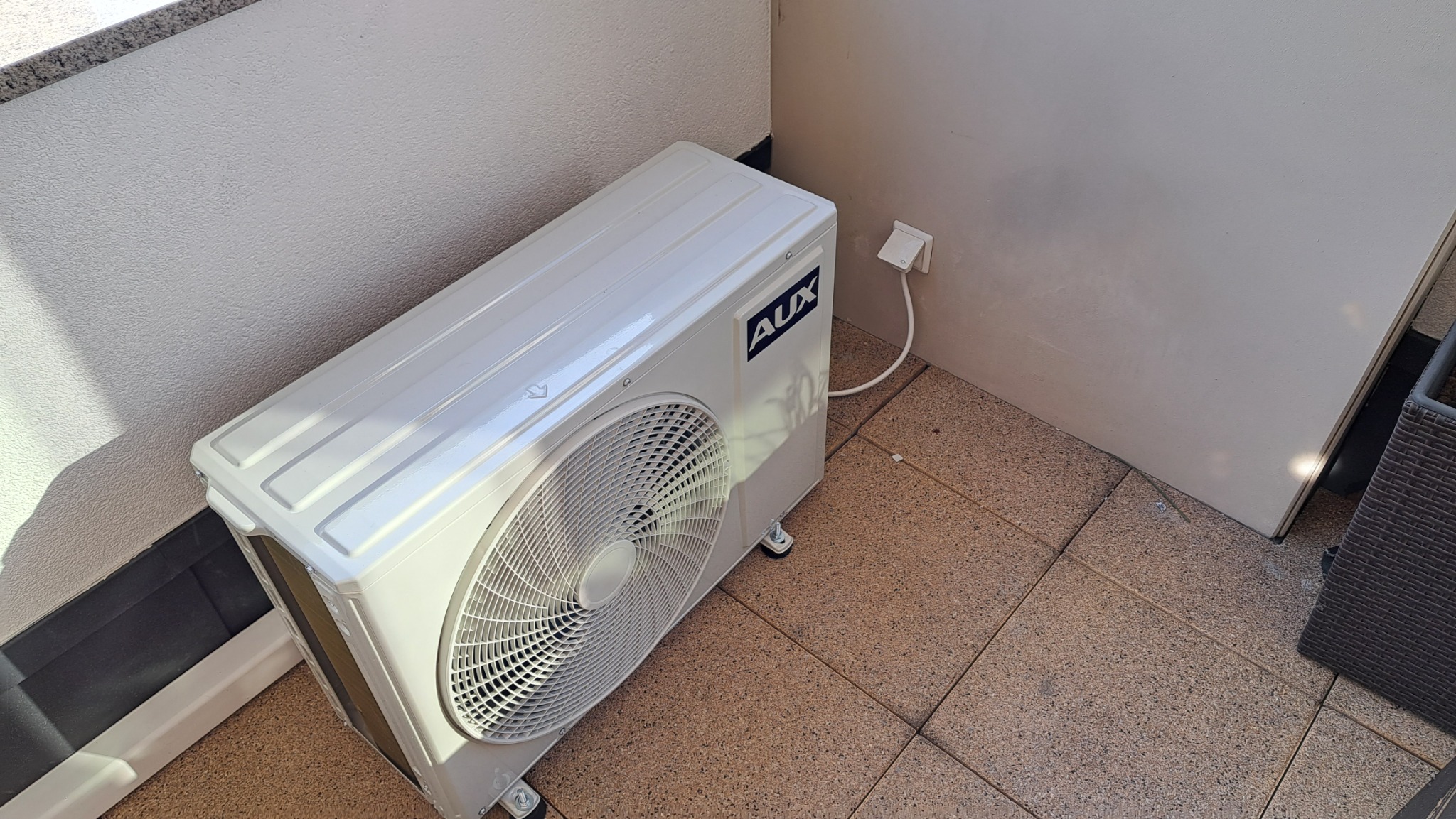 Biała jednostka zewnętrzna klimatyzatora marki AUX ustawiona na brązowym tarasie, podłączona do gniazdka elektrycznego na ścianie.