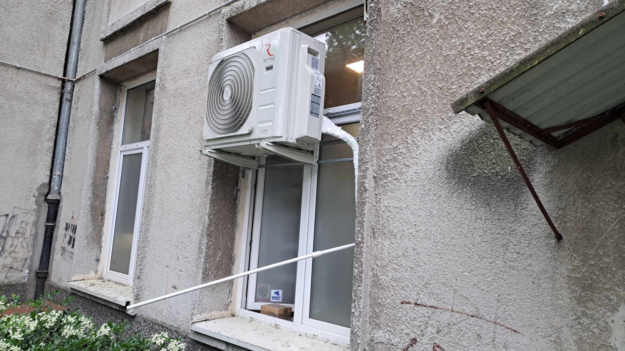 Zewnętrzna jednostka klimatyzatora Rotenso Hiro Series zamontowana na wsporniku przy oknie budynku o szarej, chropowatej elewacji.