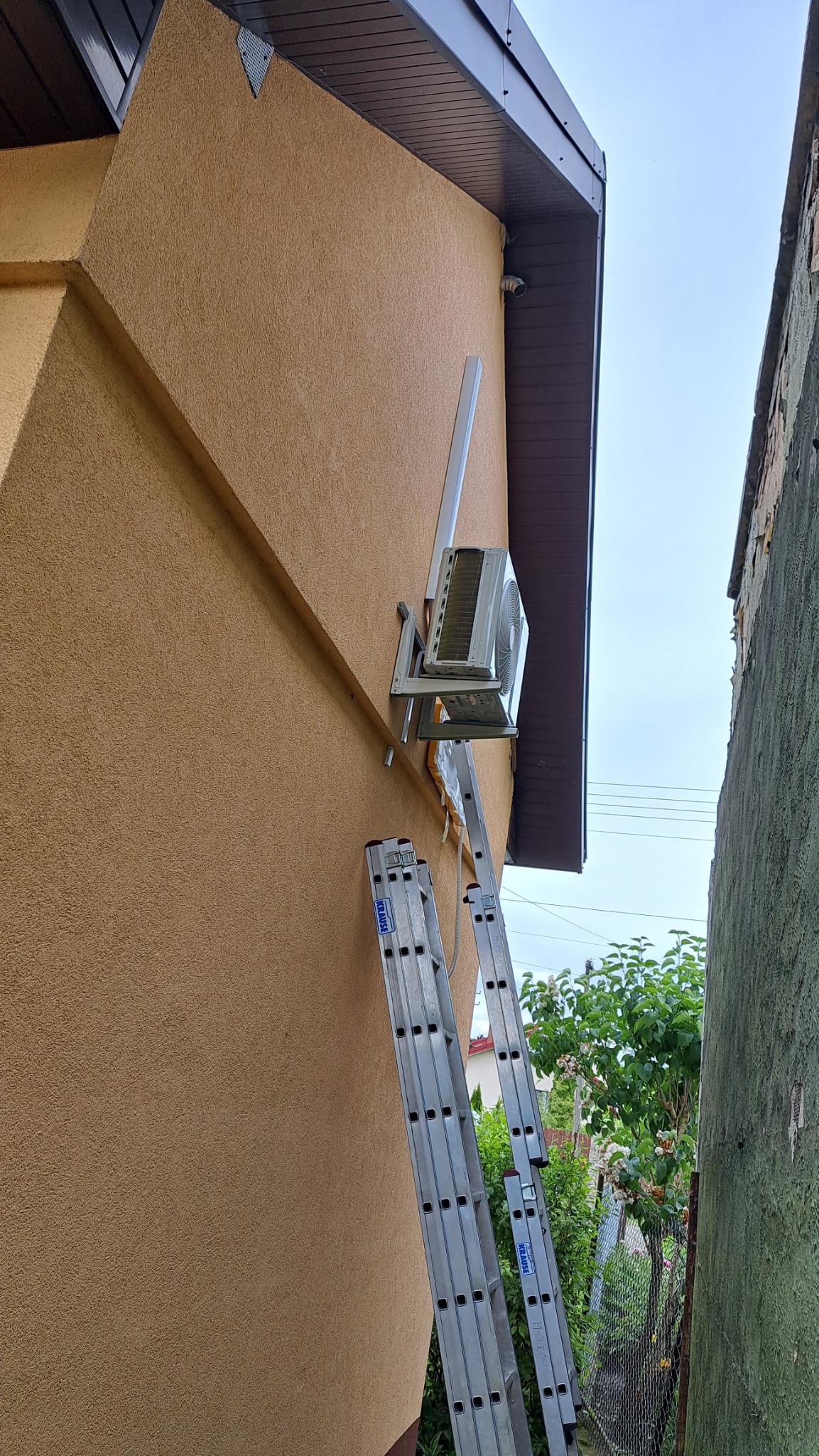 Zewnętrzna jednostka klimatyzatora zamontowana na ścianie domu, podparta drabiną Krause, widok z dołu.