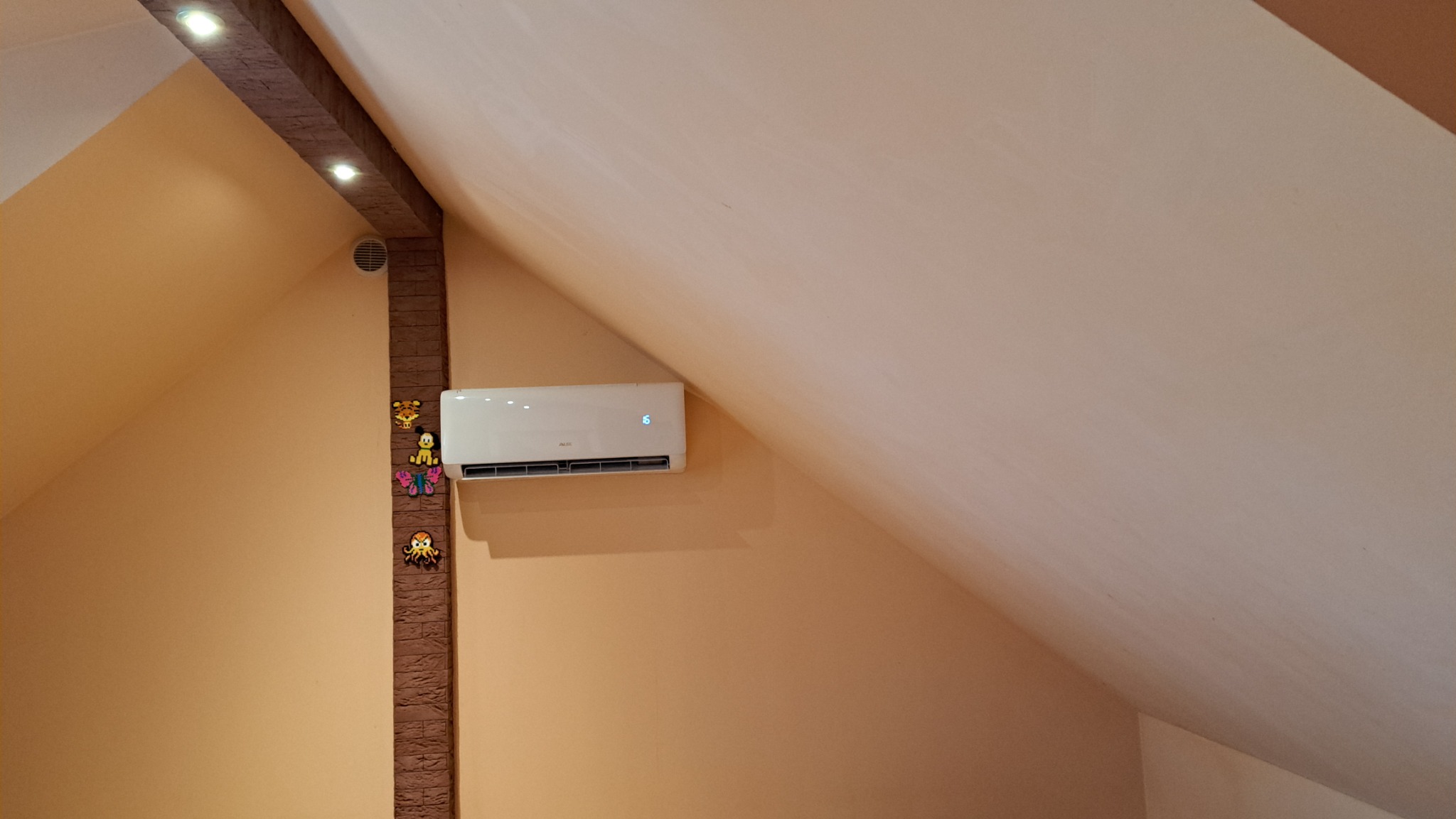 Biały klimatyzator zamontowany na skośnej ścianie poddasza, obok dekoracyjnej kolumny z oświetleniem punktowym i dziecięcymi ozdobami.