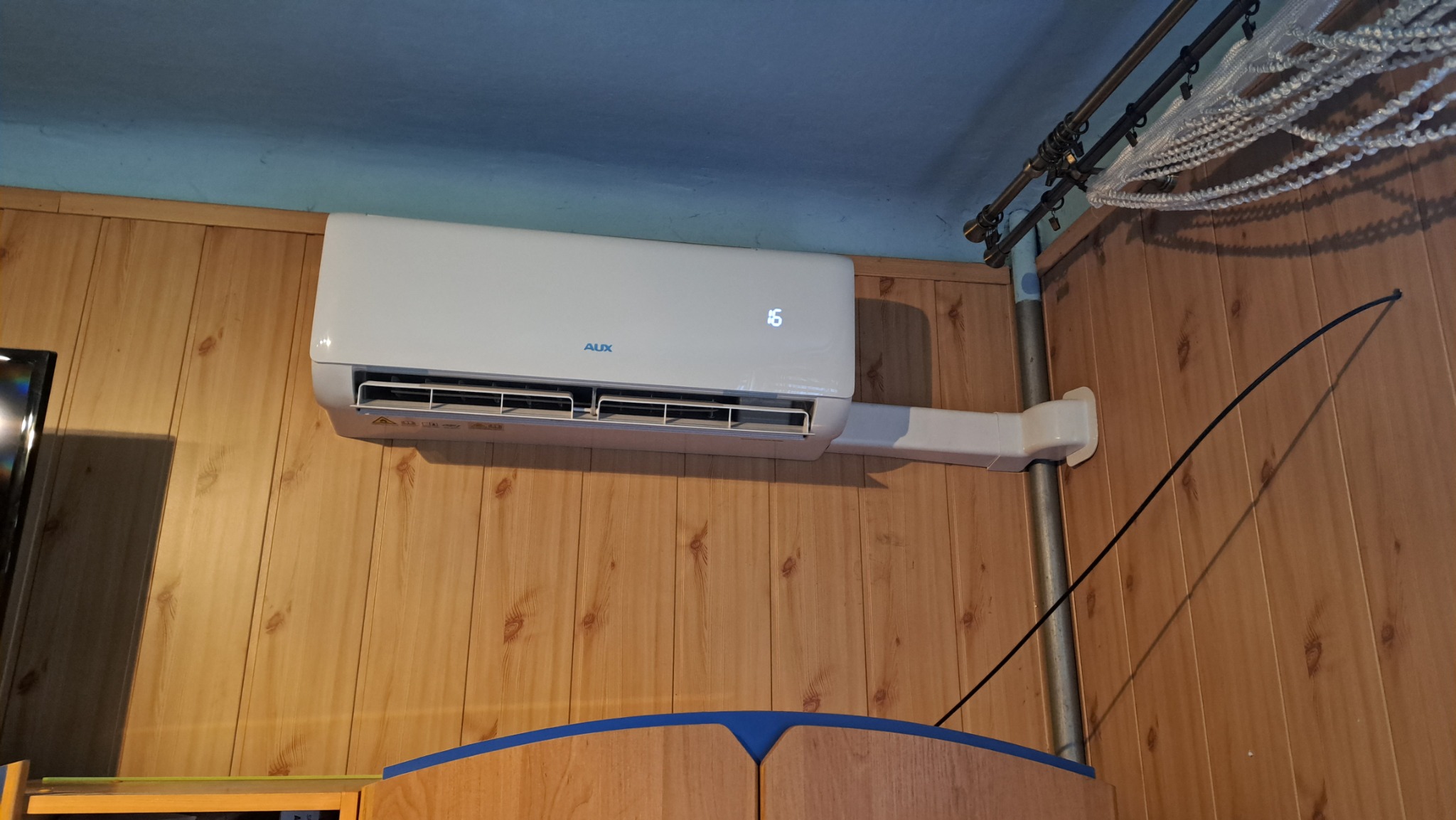 Biała jednostka klimatyzacyjna AUX zamontowana na drewnianej ścianie w pomieszczeniu z niebieskim sufitem i zasłoną z koralików.