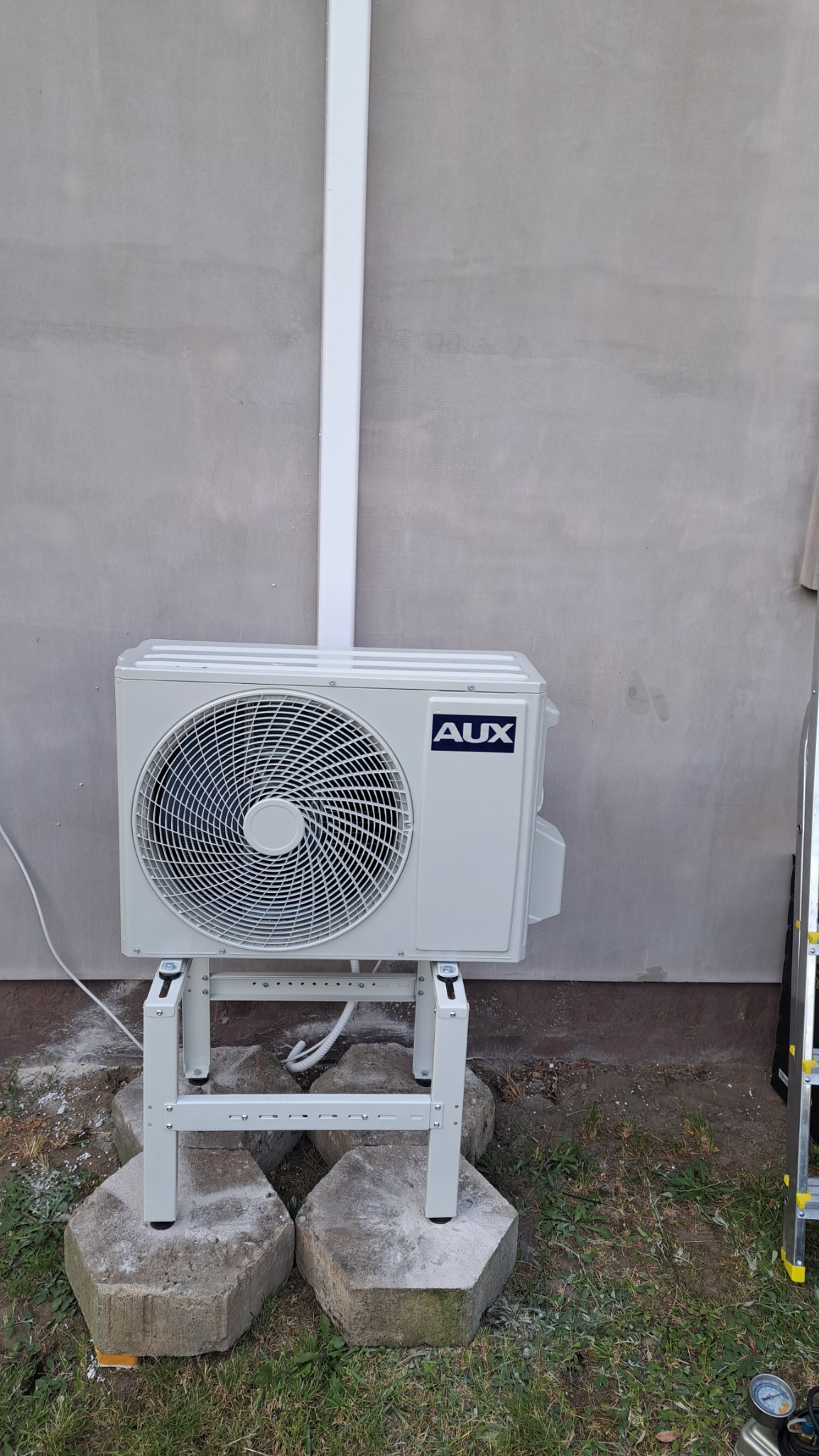 Zewnętrzna jednostka klimatyzatora marki AUX zamontowana na metalowym stelażu, stojącym na betonowych podstawach, z widocznym białym przewodem instalacyjnym biegnącym wzdłuż ściany budynku.