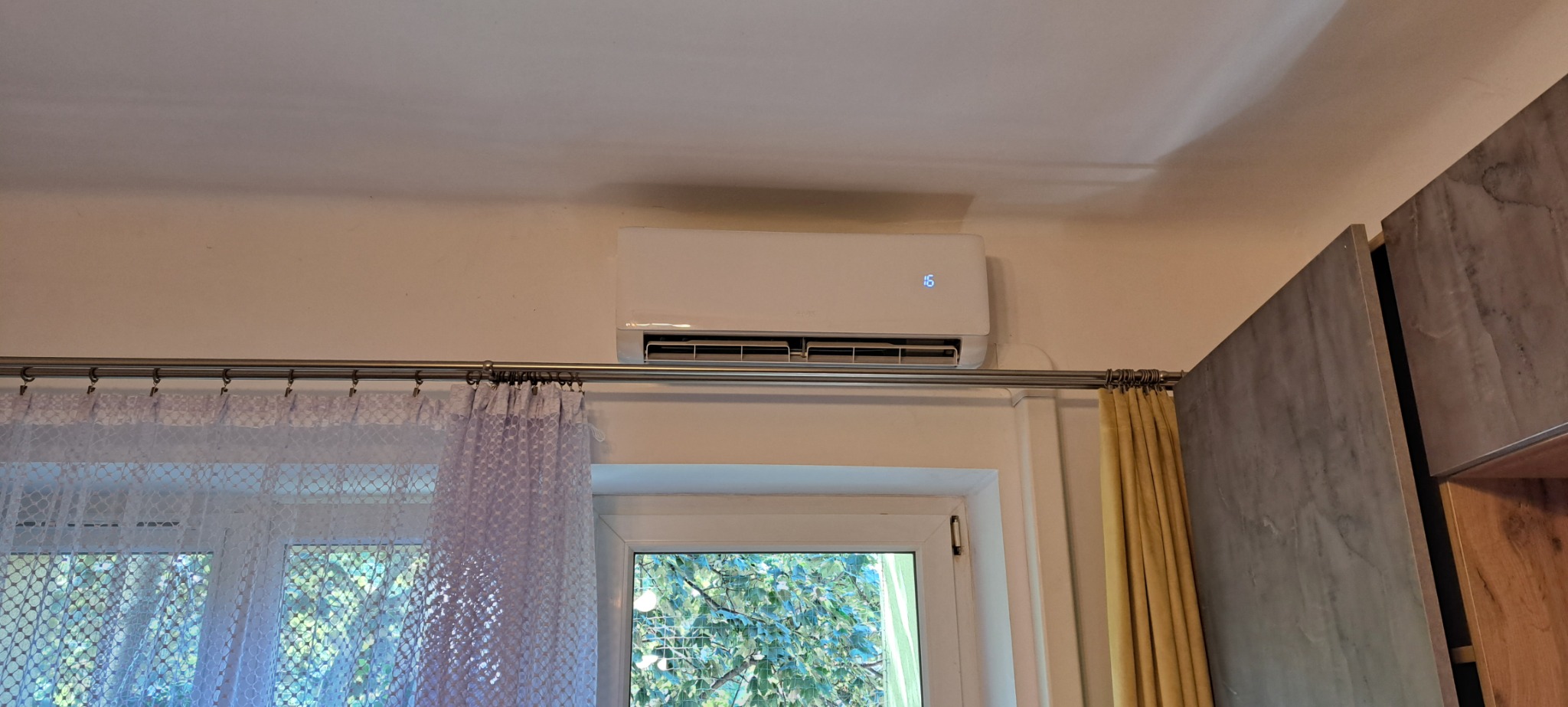 Biały klimatyzator ścienny z wyświetlaczem temperatury 16 stopni, zamontowany nad oknem z firanką i zasłoną, widok z wnętrza pomieszczenia.