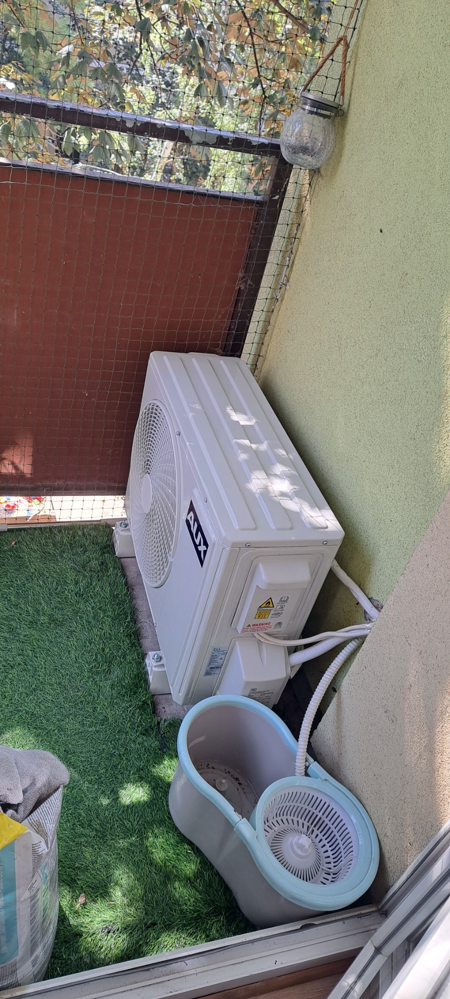 Zewnętrzna jednostka klimatyzacji AUX zamontowana na balkonie z zieloną sztuczną trawą, widoczne przewody i wiadro do mycia podłogi.