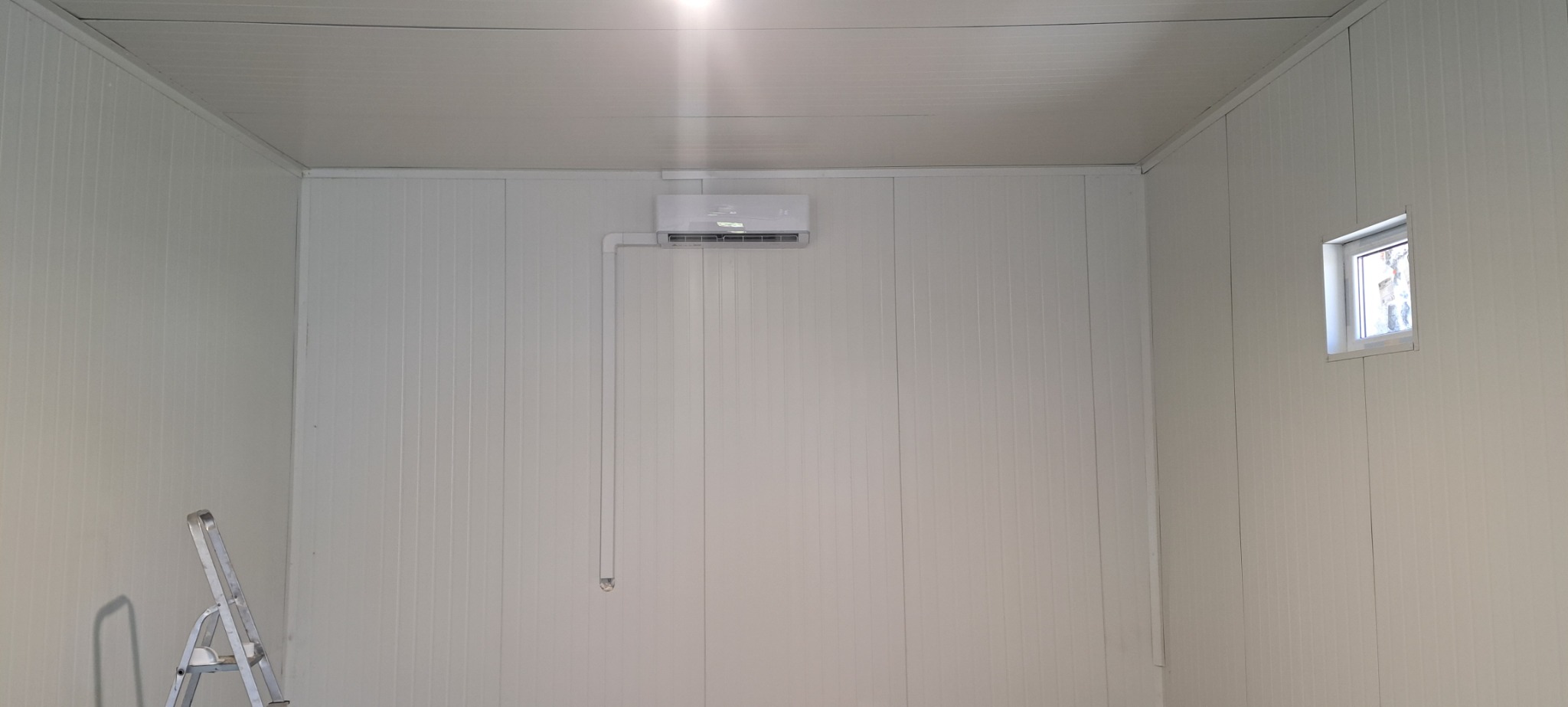Wnętrze pomieszczenia z zamontowanym klimatyzatorem na ścianie z paneli, widoczna instalacja odprowadzająca skropliny oraz drabina aluminiowa w rogu.