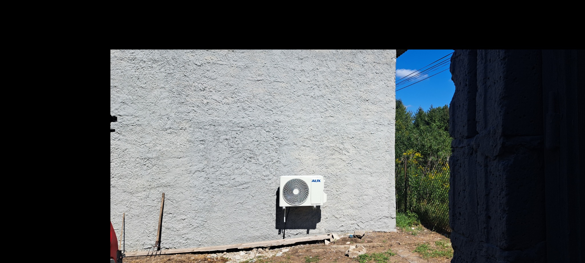 Biała jednostka zewnętrzna klimatyzacji marki AUX zamontowana na szarej, chropowatej ścianie budynku. Obok ściany widoczny fragment narzędzia ogrodniczego i drewniany element.