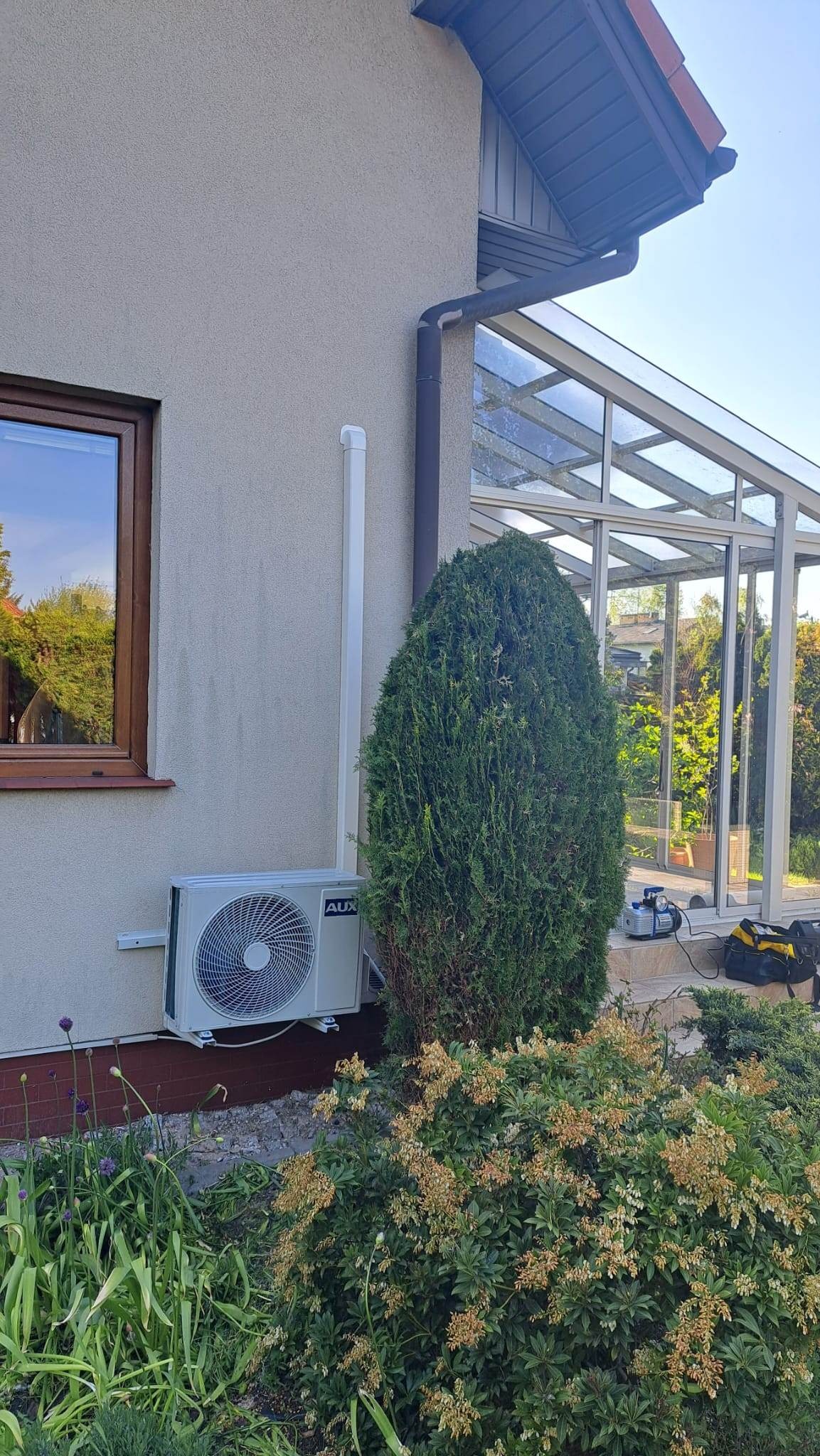 Zewnętrzna jednostka klimatyzacji AUX zamontowana na elewacji domu, obok okna i rynny, z widoczną instalacją odprowadzającą skropliny i bujną roślinnością w tle.