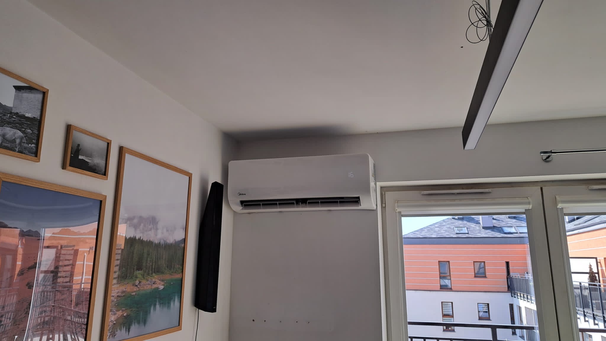 Biały klimatyzator Midea zamontowany na szarej ścianie w pokoju, widoczne ramki ze zdjęciami i okno balkonowe z widokiem na budynek.