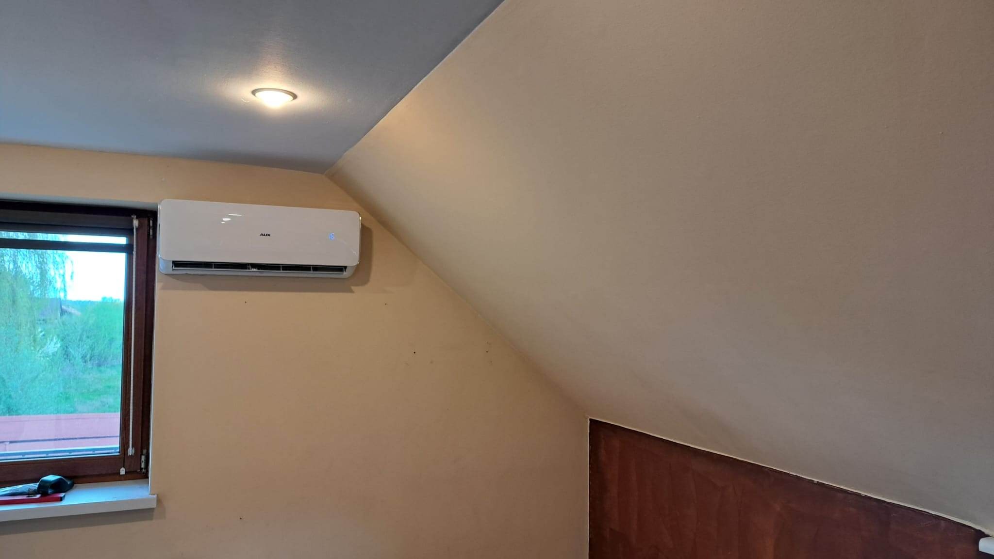 Biały klimatyzator AUX zamontowany na skośnej ścianie poddasza, widoczne okno z widokiem na zieleń.