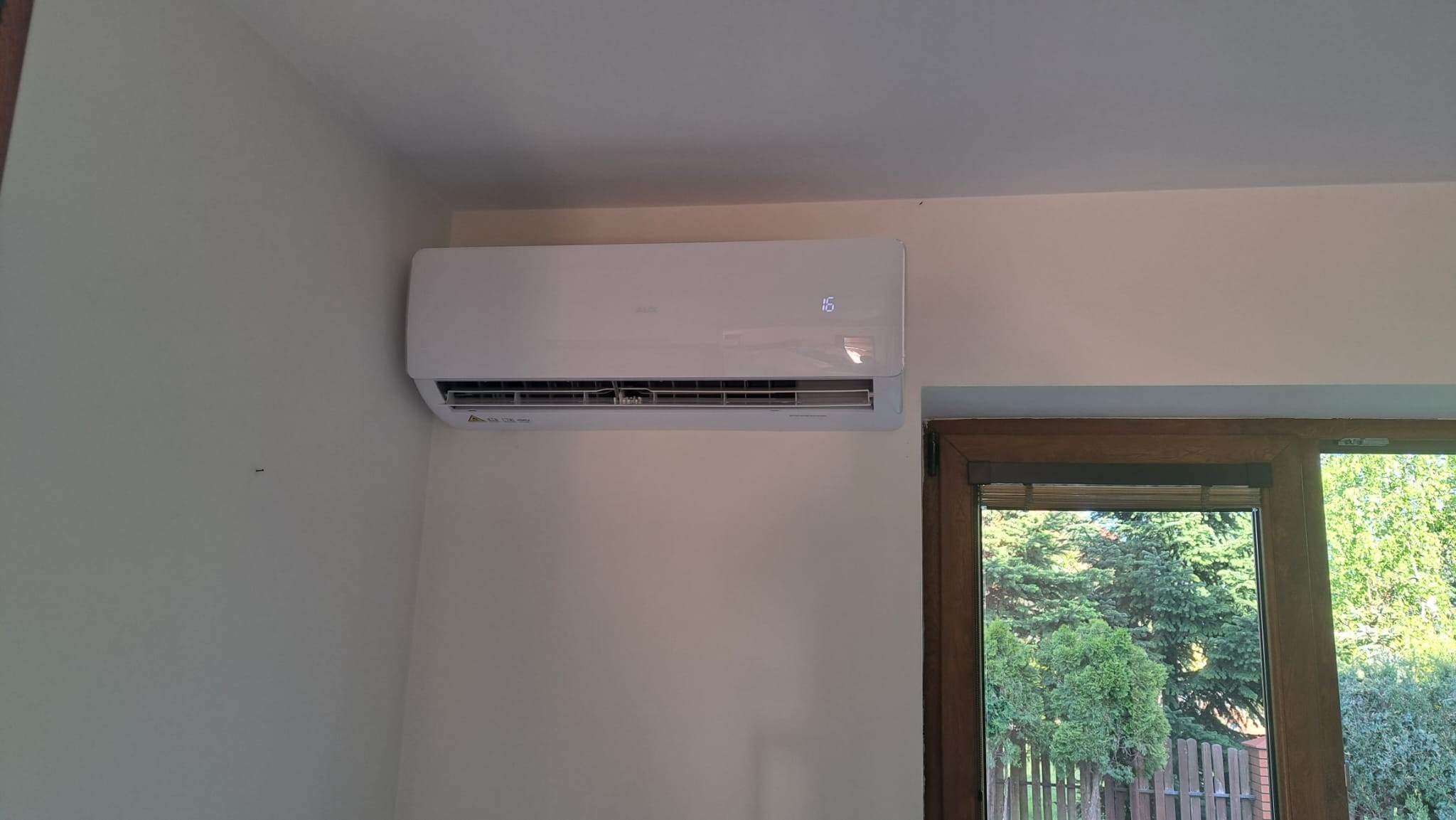 Biały klimatyzator ścienny z wyświetlaczem cyfrowym, zamontowany w jasnym pomieszczeniu obok okna z widokiem na zieleń.