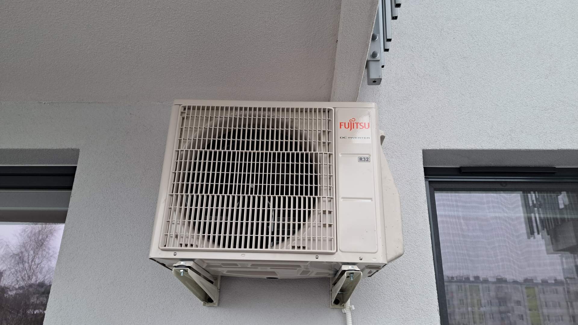 Zewnętrzna jednostka klimatyzacji Fujitsu DC Inverter R32 zamontowana na ścianie budynku, widoczna z boku okna, z elementami mocującymi.