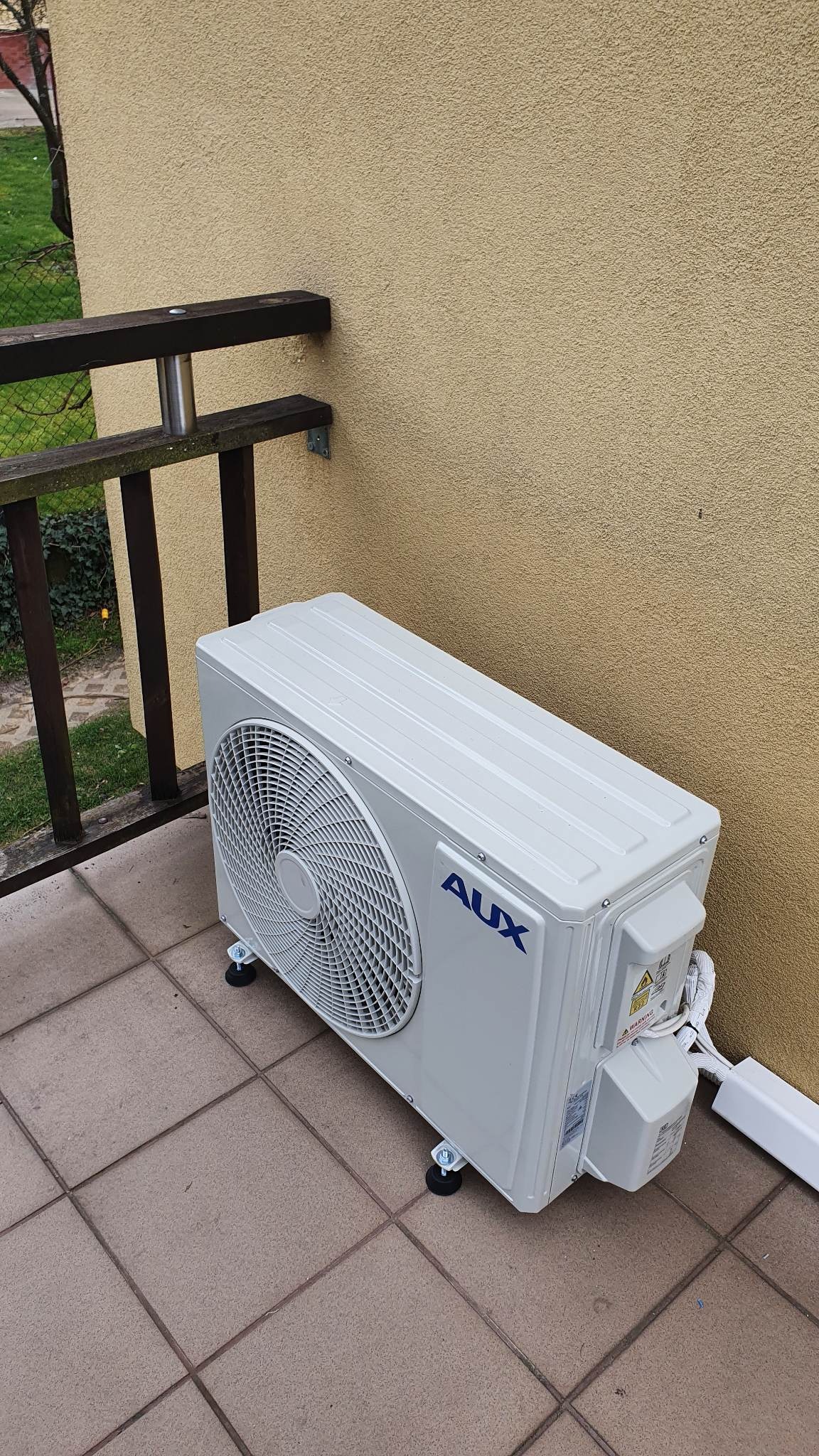 Biała jednostka zewnętrzna klimatyzacji marki AUX, umieszczona na gumowych podkładkach na brązowym tarasie przy żółtej ścianie budynku, widoczna barierka balkonu.