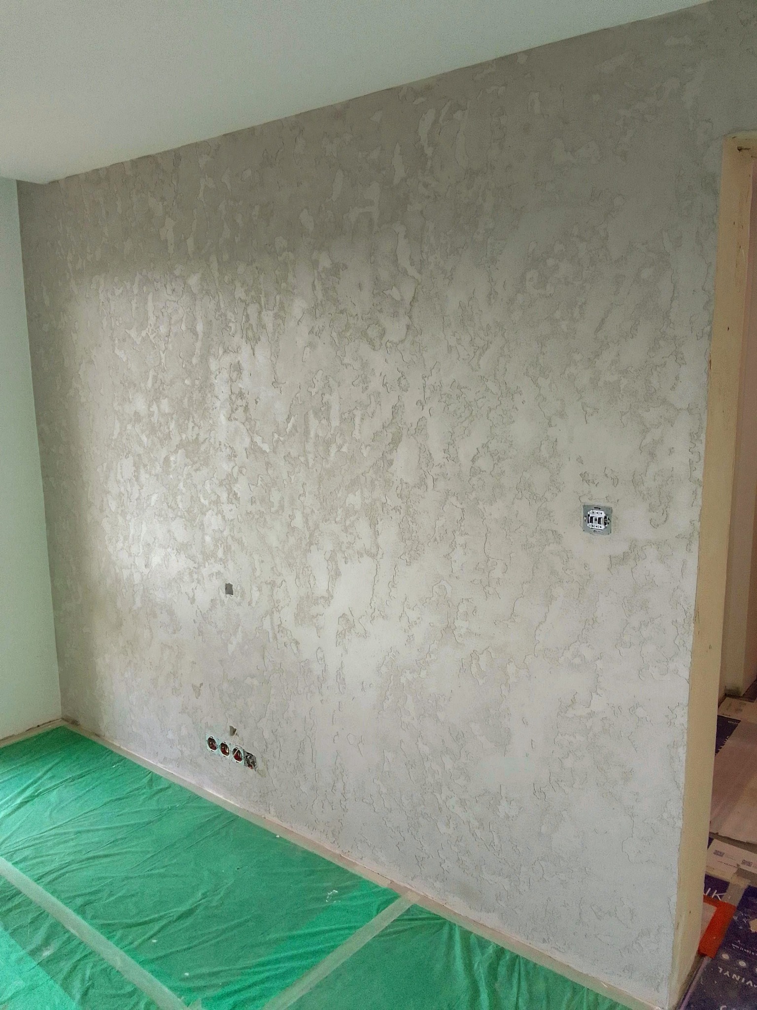 Ściana pokryta dekoracyjną, jasną strukturą, zabezpieczona podłoga folią malarską, widoczne gniazdka elektryczne i puszka instalacyjna.