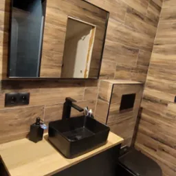 Nowoczesna łazienka z drewnopodobnymi płytkami ściennymi, czarną umywalką nablatową i toaletą w tym samym kolorze, minimalistyczne lustro z czarną ramą.