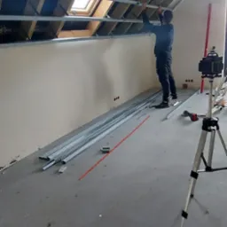 Montaż metalowego stelażu pod płytę kartonowo-gipsową na poddaszu z oknem dachowym; widoczne profile, laserowy niwelator na statywie i pracownik w trakcie mocowania.