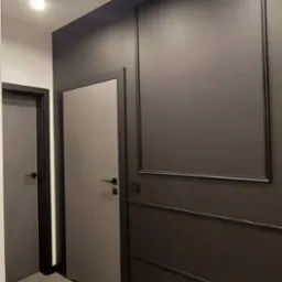 Nowoczesny korytarz w odcieniach szarości z dwiema parami drzwi o minimalistycznym designie, oświetlony trzema okrągłymi lampami sufitowymi, z dekoracyjną ścianą z panelami i ramą.