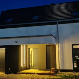 Nowoczesny dom jednorodzinny z elewacją w kolorze białym i grafitowym, oświetlony pionowymi lampami LED przy wejściu, widoczny samochód osobowy i teren wokół domu w trakcie prac wykończeniowych.