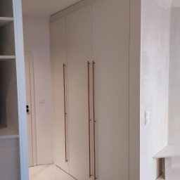 Meble Wirkos - Biała szafa wnękowa z długimi, pionowymi uchwytami w kolorze miedzi, wkomponowana w białe wnętrze z marmurową podłogą.