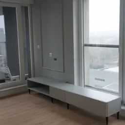 Szara szafka RTV na czarnych nóżkach, z ryflowanym frontem, ustawiona pod oknem w pustym, nowoczesnym salonie z panelami podłogowymi.