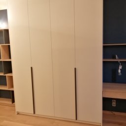 Meble Wirkos - Biała, czterodrzwiowa szafa z długimi, pionowymi uchwytami, wbudowana we wnękę z ciemnoniebieskim tłem i półkami z jasnego drewna, na podłodze z paneli.