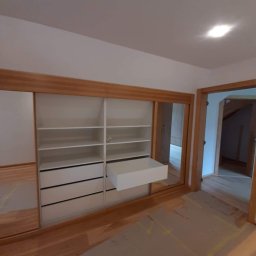 Meble Wirkos - Wnętrze z szafą wnękową z przesuwnymi lustrzanymi drzwiami, drewnianą ramą i wysuwaną białą szufladą, podłoga zabezpieczona kartonem.