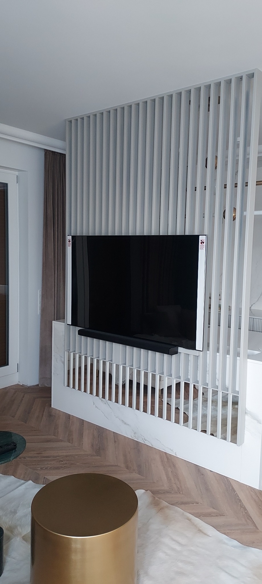 Nowoczesna aranżacja salonu z telewizorem na ścianie z białych, pionowych listew, marmurowy cokół, parkiet w jodełkę i złoty stolik kawowy