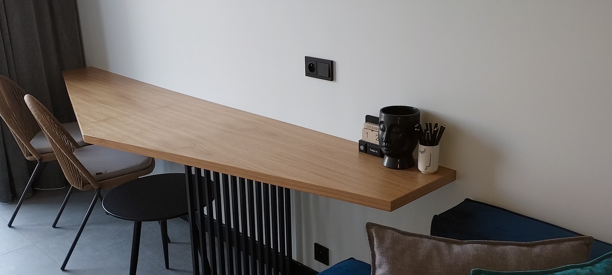 Długi, wąski blat drewniany zamontowany do ściany, wsparty na czarnych, pionowych listwach, z dwoma krzesłami obok i czarną lampą w kształcie głowy oraz kalendarzem i pojemnikiem z długopisami...