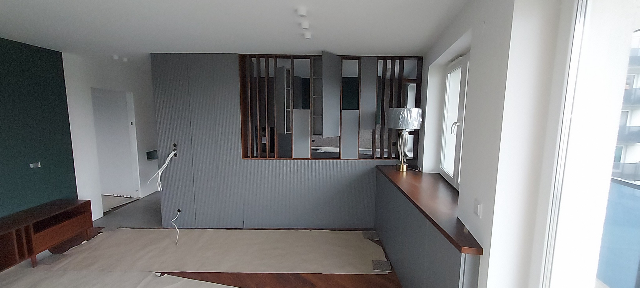 Szara zabudowa meblowa z lamelami i otwartymi półkami w trakcie montażu, widoczne kable instalacyjne, zabezpieczona podłoga.