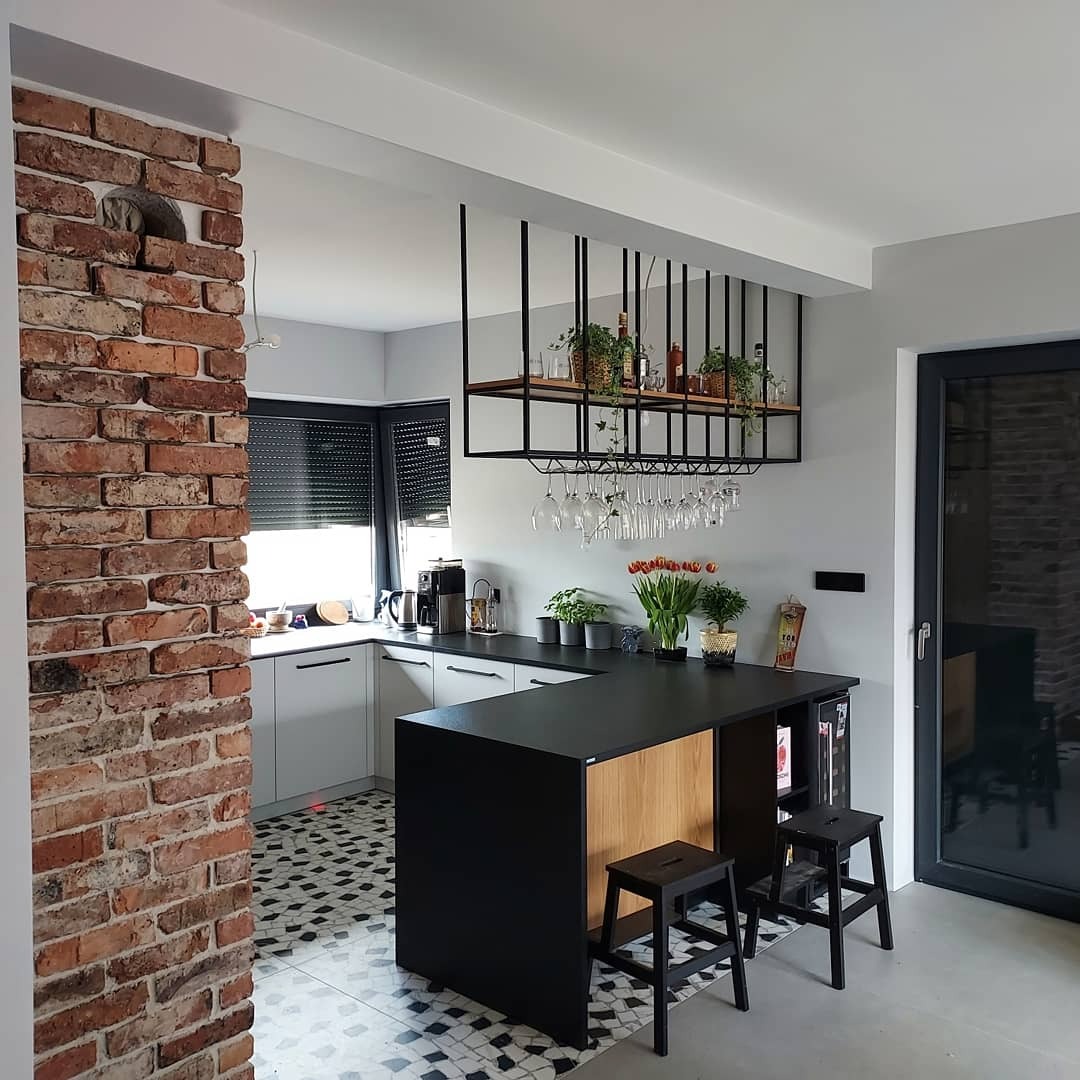 Nowoczesna kuchnia z wyspą, czarnym blatem, ceglaną ścianą i wiszącym regałem na kieliszki oraz rośliny.