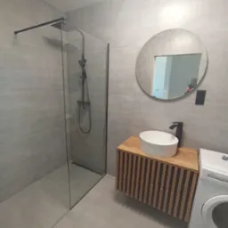 Nowoczesna łazienka z szarą glazurą, kabiną prysznicową typu walk-in z czarną armaturą, okrągłym lustrem i drewnianą szafką pod umywalką oraz pralką.