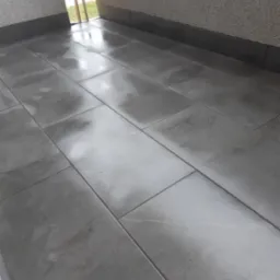 Szare, prostokątne płytki podłogowe z delikatnym wzorem marmuru, ułożone wzdłuż ściany z szorstką fakturą, widoczne okno z widokiem na zieleń na zewnątrz.