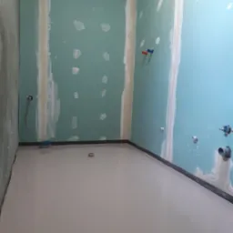 Surowy stan łazienki w trakcie remontu: ściany z płyt karton gips, częściowo zaszpachlowane, przygotowane do malowania, wylewka podłogowa, wystające rury instalacji wodnej i kanalizacyjnej.