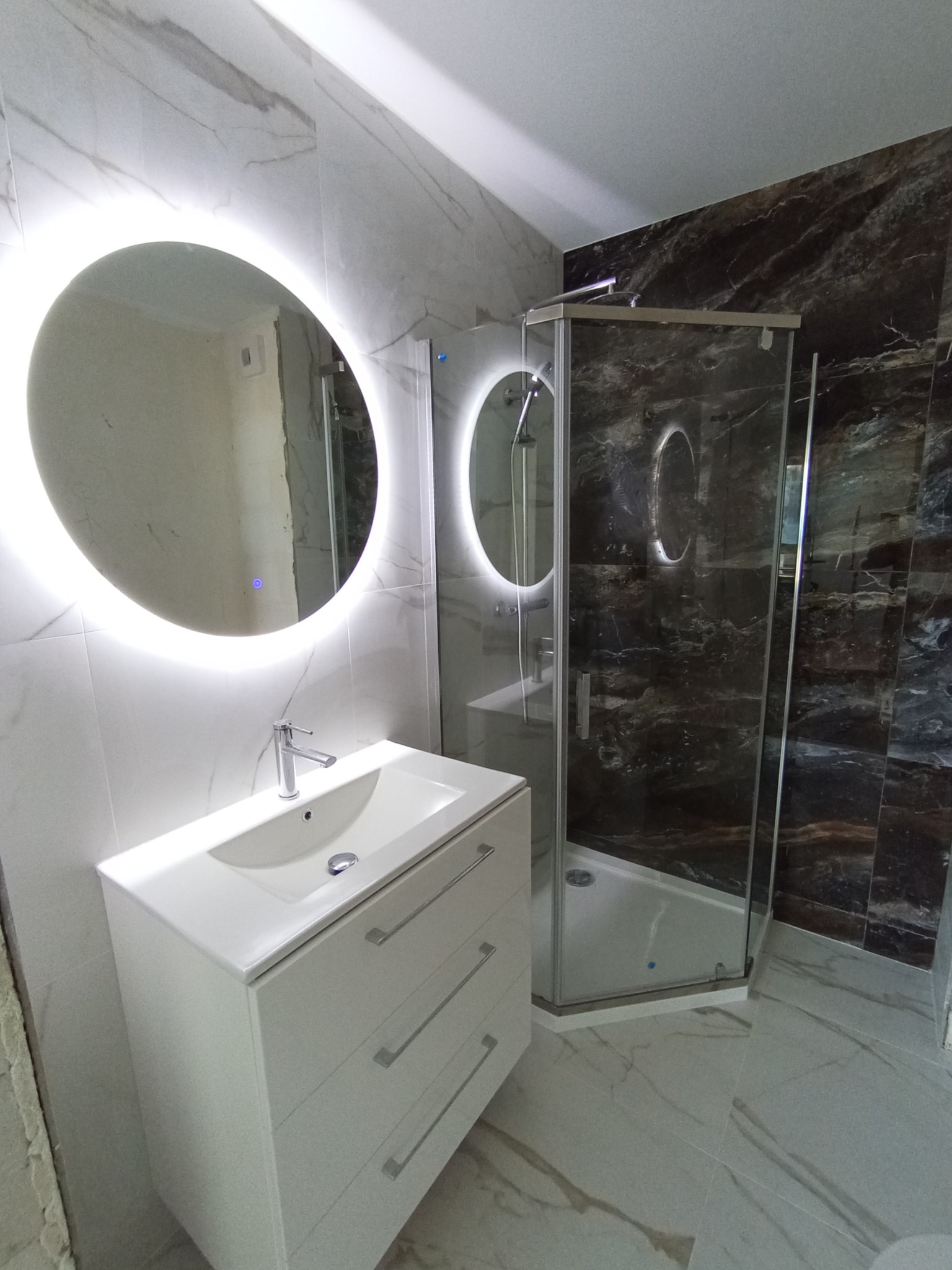 Nowoczesna łazienka z okrągłym lustrem LED, białą szafką z umywalką i kabiną prysznicową z ciemnymi płytkami imitującymi marmur.