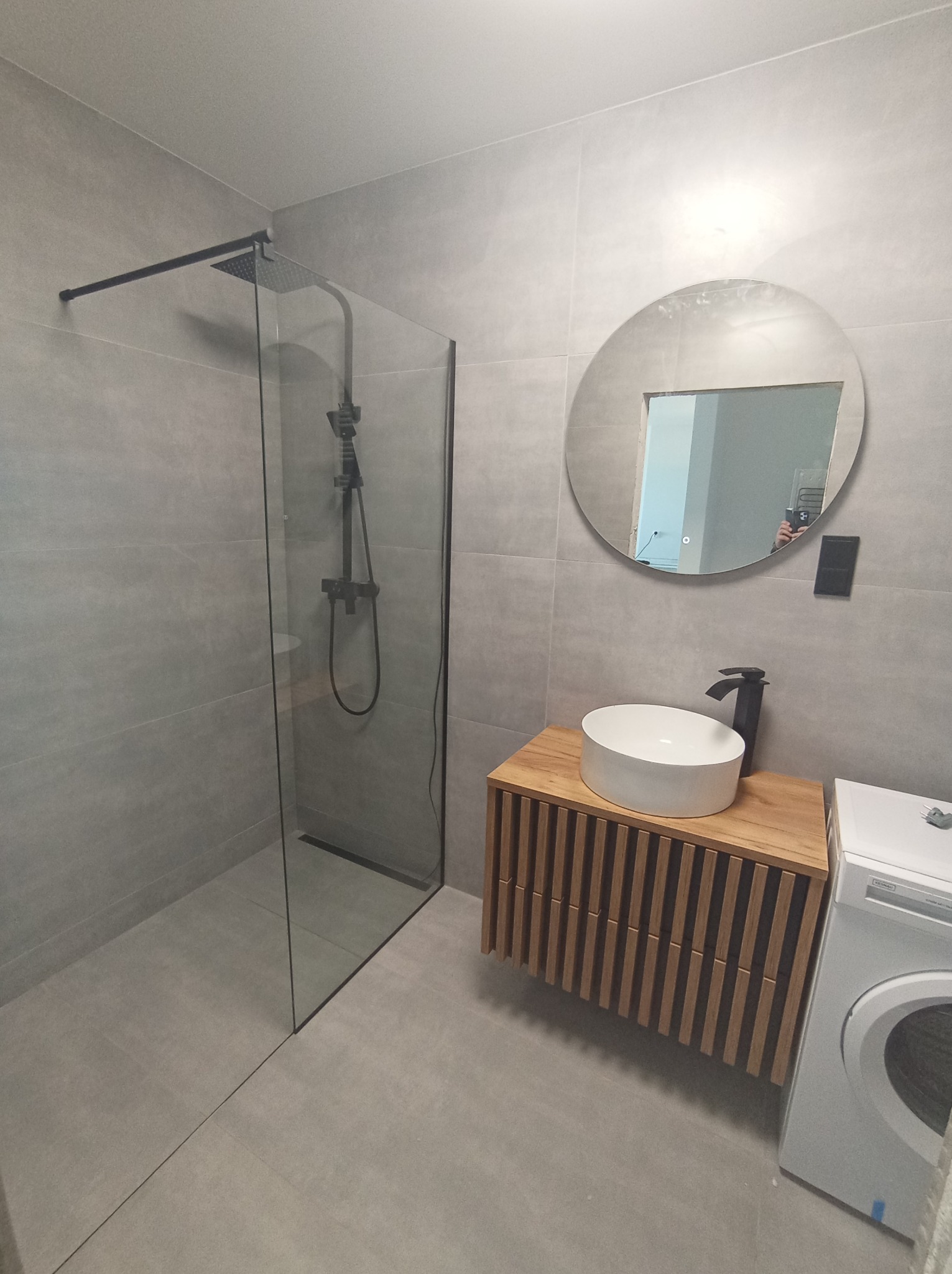 Nowoczesna łazienka z szarą glazurą, kabiną prysznicową typu walk-in z czarną armaturą, okrągłym lustrem i drewnianą szafką pod umywalką oraz pralką.