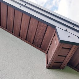 DMX USŁUGI TRANSPORTOWO - BUDOWLANE Jarosław Wolski - Detal wykończenia elewacji domu: brązowa podbitka dachowa z widoczną strukturą drewna, szara listwa okapowa i jasny tynk strukturalny na ścianie. Ujęcie z dołu.