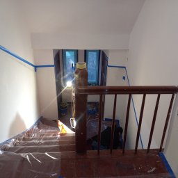 DMX USŁUGI TRANSPORTOWO - BUDOWLANE Jarosław Wolski - Klatka schodowa zabezpieczona folią i taśmą malarską przed malowaniem ścian. Widoczne narzędzia i oświetlenie. Ujęcie z góry, zza drewnianej balustrady.