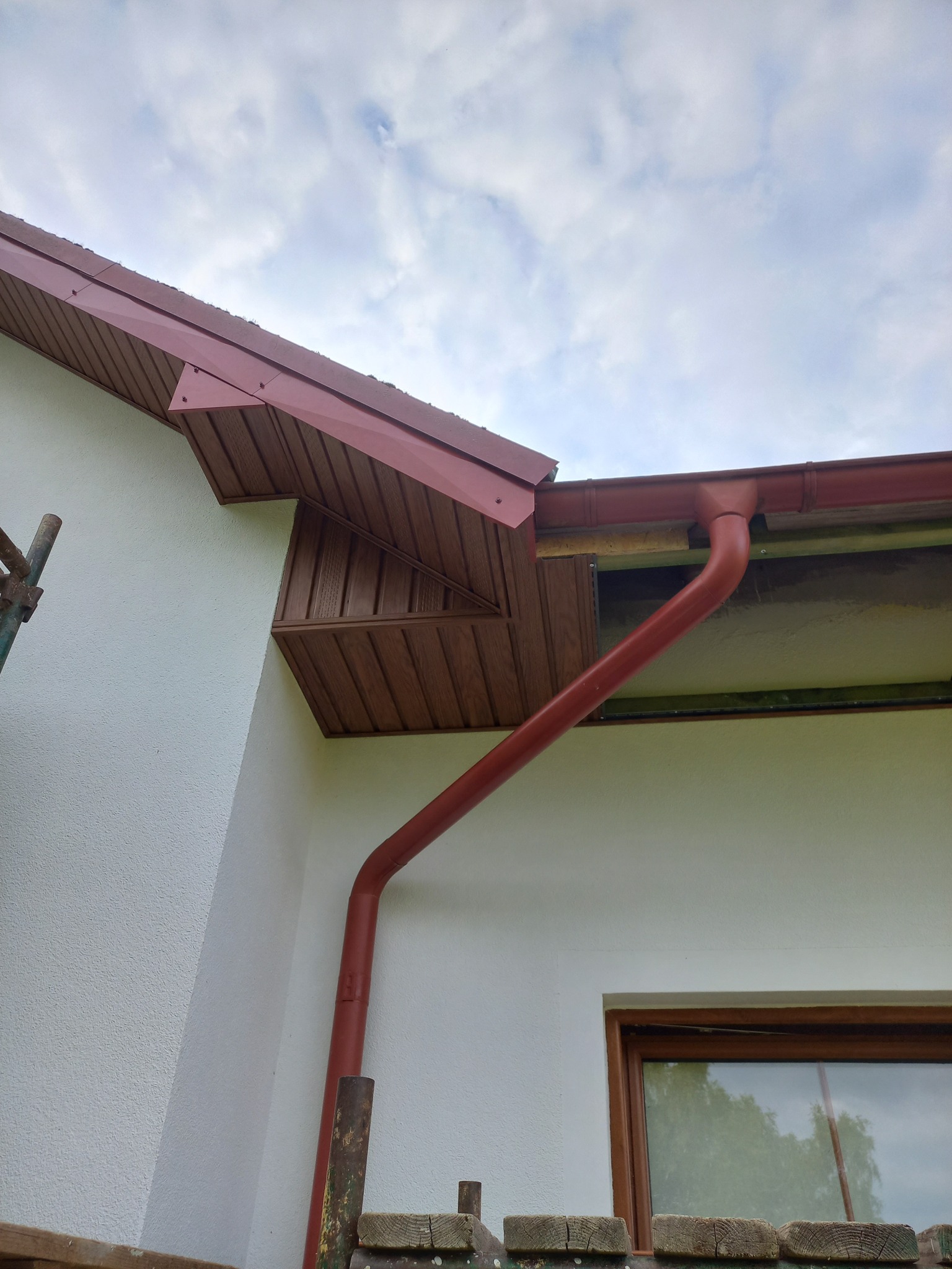 Fragment elewacji domu z brązowym orynnowaniem i podbitką. Widoczne okno i fragment rusztowania. Teksturowana, biała ściana i pochmurne niebo w tle.