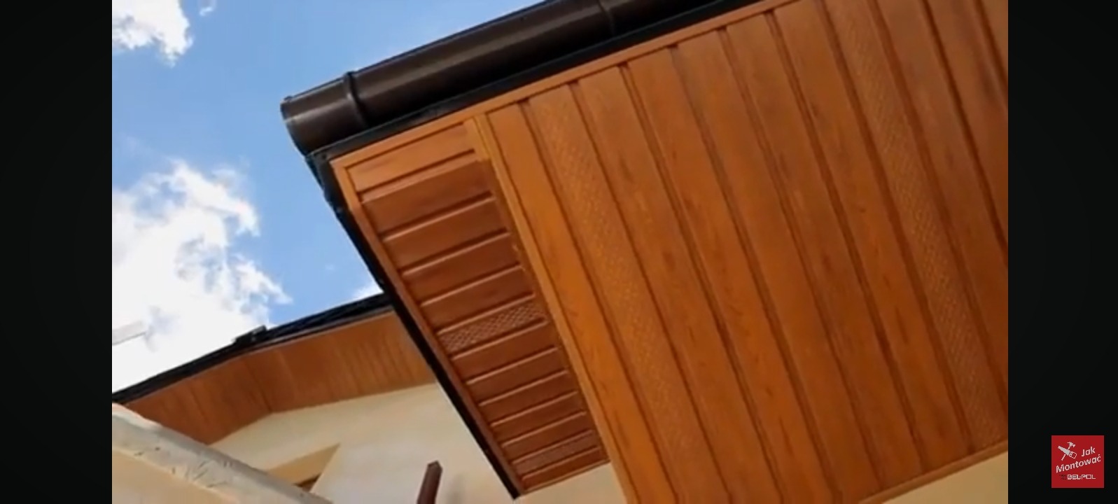 Drewniana podbitka dachowa w odcieniach brązu, widoczna z dołu pod kątem. Na górze brązowa rynna, w tle fragment błękitnego nieba z chmurami.