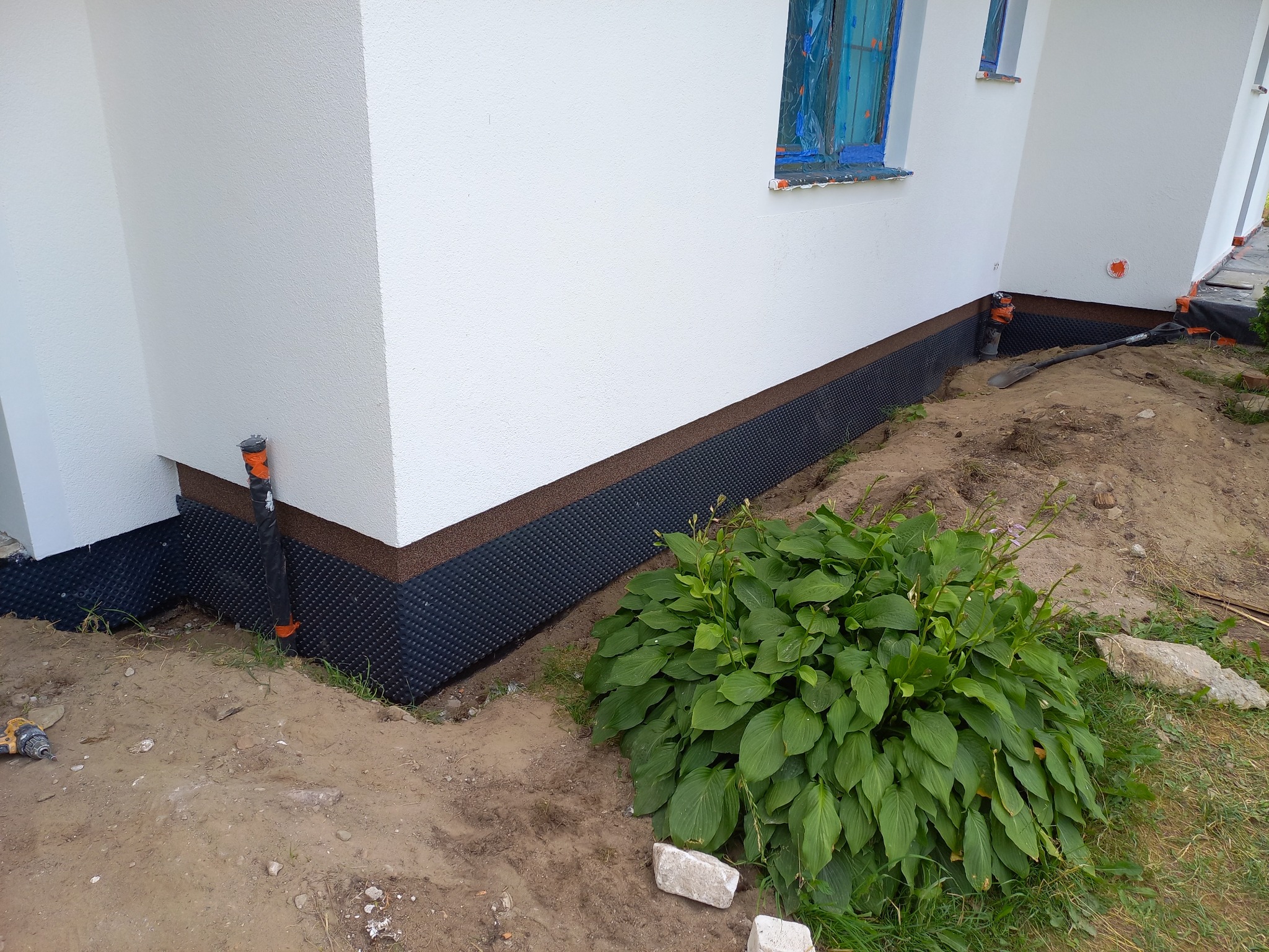 Ocieplona, biała elewacja budynku z widoczną hydroizolacją fundamentów i zabezpieczonymi oknami folią. W tle ziemia i roślinność.