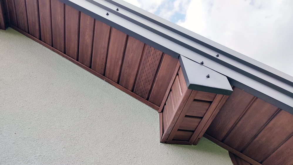 Detal wykończenia elewacji domu: brązowa podbitka dachowa z widoczną strukturą drewna, szara listwa okapowa i jasny tynk strukturalny na ścianie. Ujęcie z dołu.