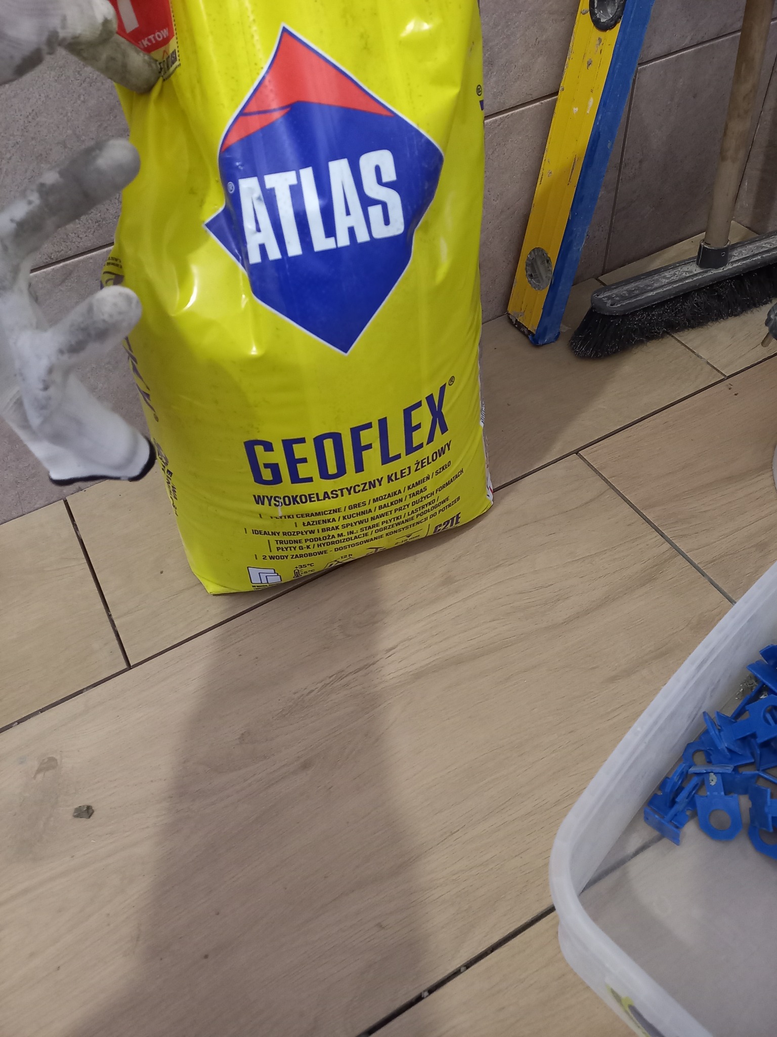 Żółty worek kleju Atlas Geoflex na podłodze z płytek, obok poziomica, szczotka i niebieskie krzyżyki dystansowe. Widoczne rękawice robocze.