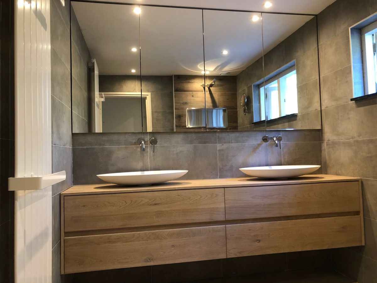 Nowoczesna łazienka z drewnianą szafką pod umywalką i dużym lustrem. Szare płytki na ścianach i podłodze. Minimalistyczny design, oświetlenie punktowe.