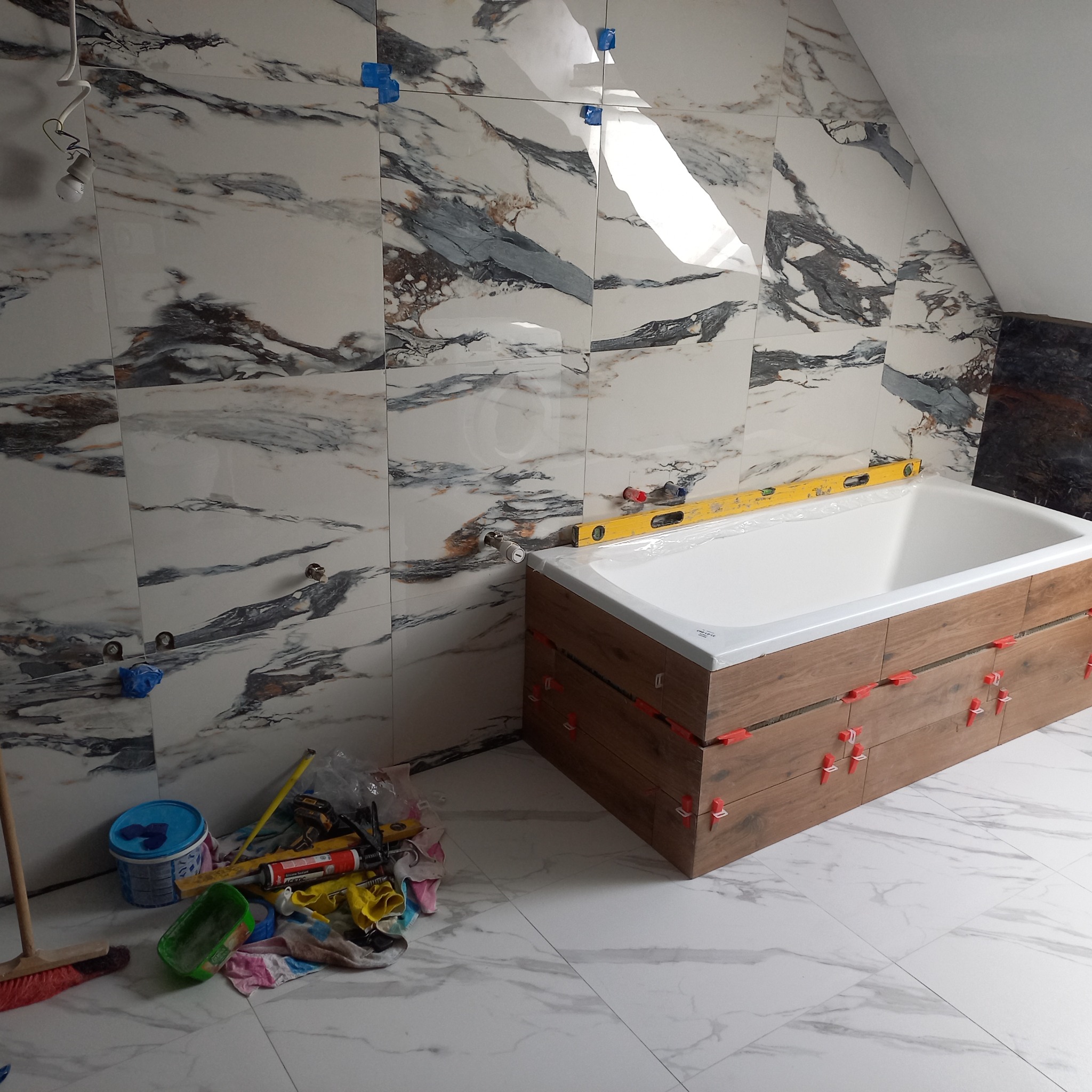 Łazienka w trakcie wykańczania: marmurowe płytki na ścianach z poziomnicą nad wanną obudowaną drewnopodobnymi płytkami. Widoczne narzędzia i akcesoria glazurnicze.
