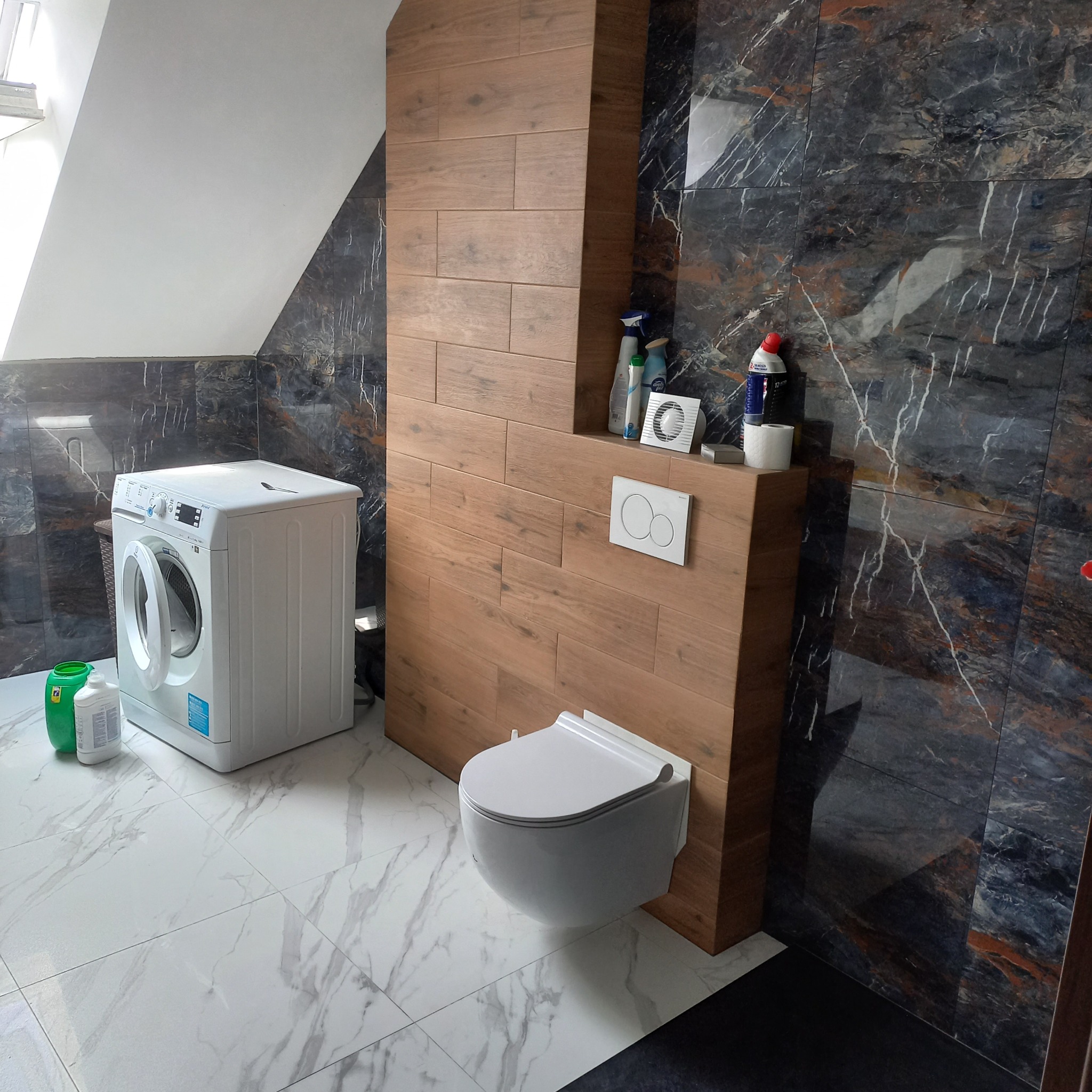 Nowoczesna łazienka na poddaszu z marmurowymi płytkami, drewnopodobną zabudową WC i pralką. Białe płytki podłogowe z szarym wzorem żyłkowania.